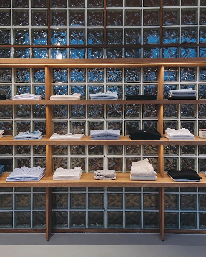 Organized display of folded shirts on wooden shelves against a glass block wall.